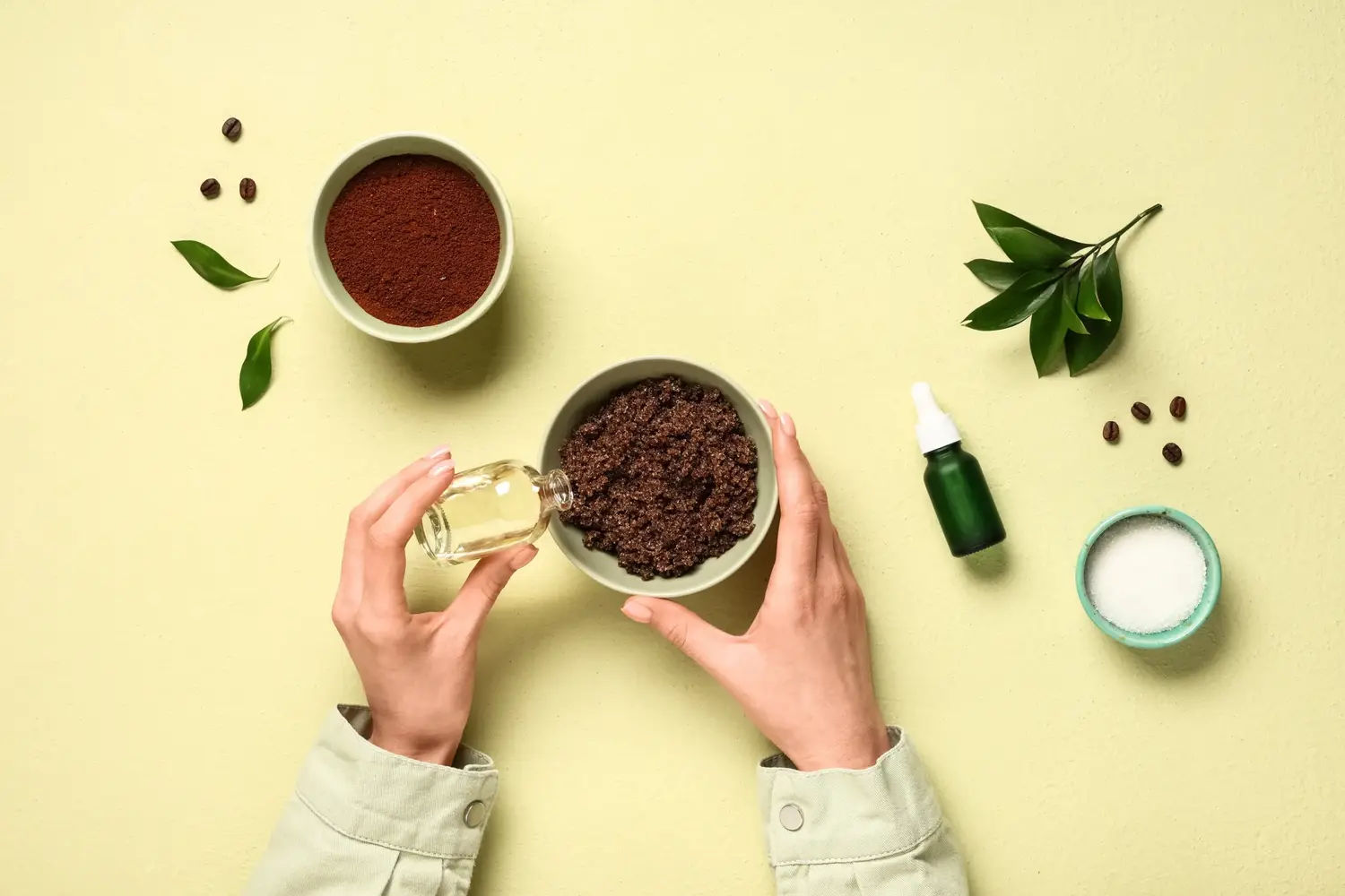 Immagine dall'alto di un ciotola con scrub corpo al caffè, foglie e chicchi con due mani in primo piano